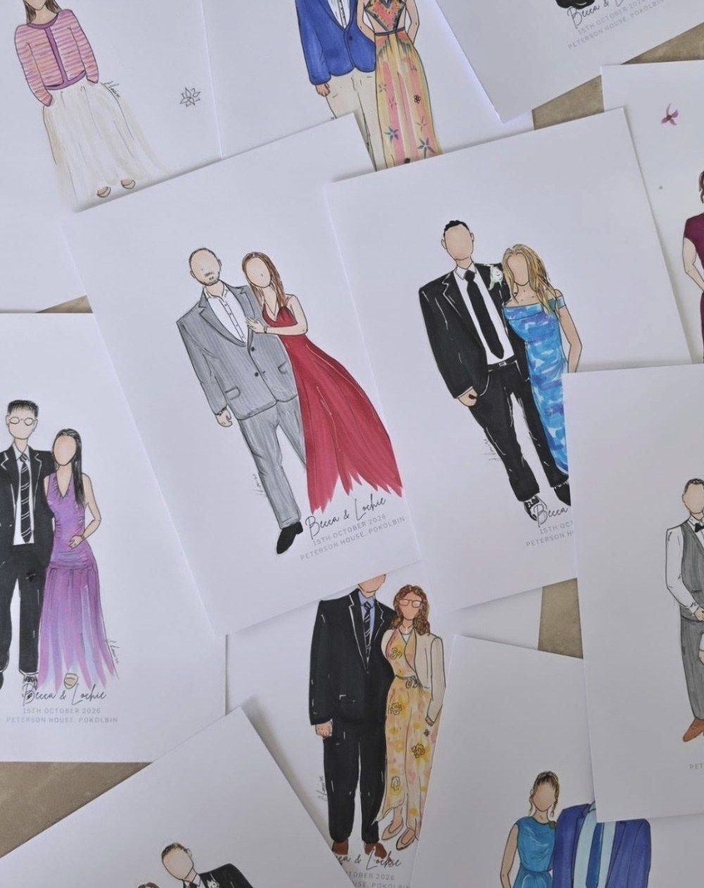 A table covered in finished keepsake sketches from one wedding, each guest coloured in and lettered with the couple's names, date and venue